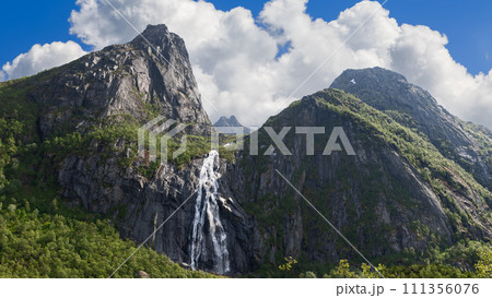 Sun-dappled Valefossen waterfall cascades down Lofoten's cliffs, lush greenery, dramatic peaks 111356076