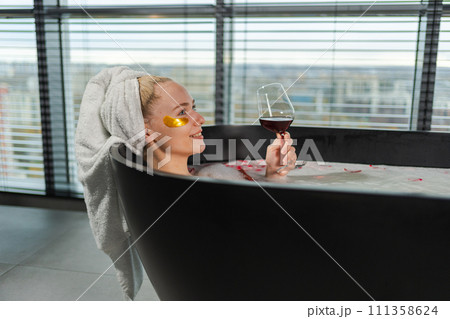 Spa relaxation. Woman lying in bath with holding glass with red wine. Girl relaxing in bathroom at home. Pretty female taking hot bath drinking wine. Stress relief. Rest after hard working day 111358624