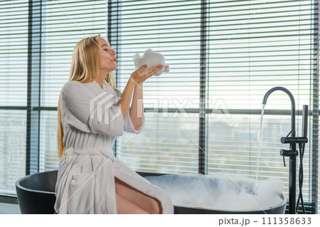 Spa relaxation. Young woman in spa bathrobe sitting at bath blowing foam in bathtub. Girl relaxing in bathroom at home. Female spending morning in bathroom. Beauty treatment body skin care concept 111358633