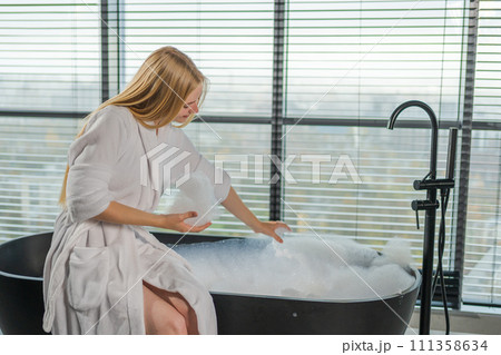 Spa relaxation. Young woman in spa bathrobe sitting at bath with foam in bathtub. Girl relaxing in bathroom at home. Female spending morning in bathroom. Beauty treatment body skin care concept 111358634