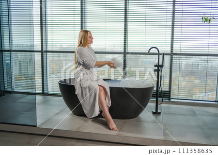 Spa relaxation. Young woman in spa bathrobe sitting at bath with foam in bathtub. Girl relaxing in bathroom at home. Female spending morning in bathroom. Beauty treatment body skin care concept Spa relaxation. Young woman in spa bathrobe sitting at bath with foam in bathtub. Girl relaxing in bathroom at home. Female spending morning in bathroom. Beauty treatment body skin care concept 111358635