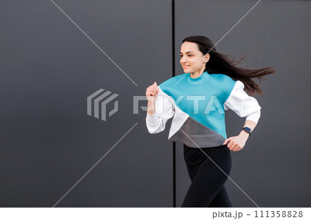 Young woman with fit body jumping and running against dark wall background. Female model in sportswear exercising outdoors. Young woman with fit body jumping and running against dark wall background. Female model in sportswear exercising outdoors. 111358828