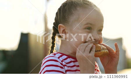 Funny cute little girl fooling eating big piece of fast food burger cheeseburger hamburger enjoy happy childhood. Cheerful female kid child biting junk meal with bun and meat bright sun light outdoor 111360991