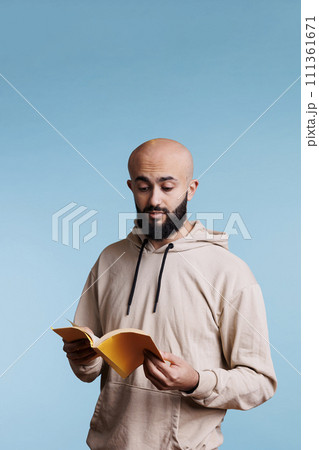 Arab man reading book paperback edition with yellow cover. Young concentrated bald bearded person in hoodie holding open notepad while standing with surprised facial expression Arab man reading book paperback edition with yellow cover. Young concentrated bald bearded person in hoodie holding open notepad while standing with surprised facial expression 111361671