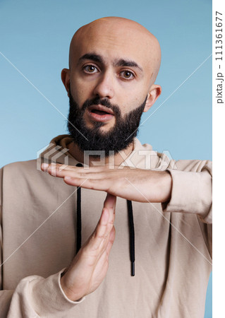 Arabian person taking time out with stop signal while showing confident facial expression portrait. Young bald bearded man making interruption gesture with hands while looking at camera 111361677