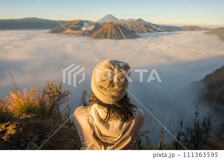 Back view of Asian women standing on the hill and looking to the beautiful sunrise and sea of fog over Bromo Tengger Semeru National Park in East Java, Indonesia. Back view of Asian women standing on the hill and looking to the beautiful sunrise and sea of fog over Bromo Tengger Semeru National Park in East Java, Indonesia. 111363595