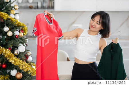 Happy young Asian girl trying to choose a dress standing next to Xmas-tree 111364262