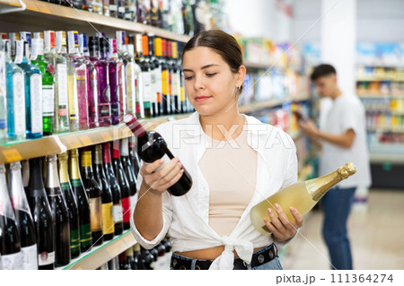 in supermarket young woman carefully chooses wine. in supermarket young woman carefully chooses wine. 111364274