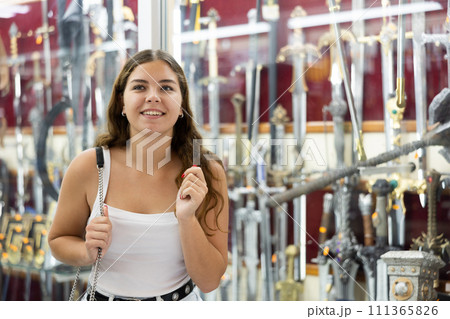 Interested young girl looking at crafted swords in souvenir shop Interested young girl looking at crafted swords in souvenir shop 111365826
