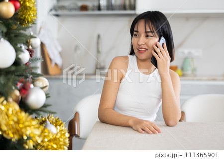 Smiling young Asian girl speaking on the phone sitting at table by Xmas tree 111365901