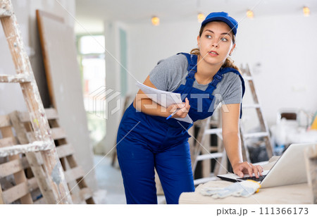 Portrait of a female builder who checks the work done using laptop Portrait of a female builder who checks the work done using laptop 111366173