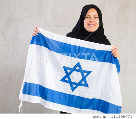 Balanced smiling Muslim woman in traditional black hijab holds flag of Israel 111366475
