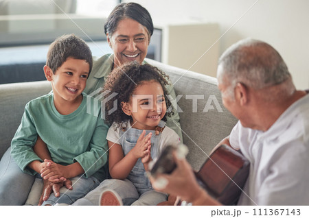Grandfather playing the guitar for grandmother and children sitting on the sofa in their home. Retired grandparents entertain happy and smiling grandchildren with music on acoustic guitar and bonding Grandfather playing the guitar for grandmother and children sitting on the sofa in their home. Retired grandparents entertain happy and smiling grandchildren with music on acoustic guitar and bonding 111367143