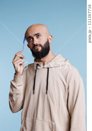 Pensive arab man holding pen to head while thinking and looking away. Young arabian person wearing casual hoodie making decision and remembering with thoughtful expression Pensive arab man holding pen to head while thinking and looking away. Young arabian person wearing casual hoodie making decision and remembering with thoughtful expression 111367716
