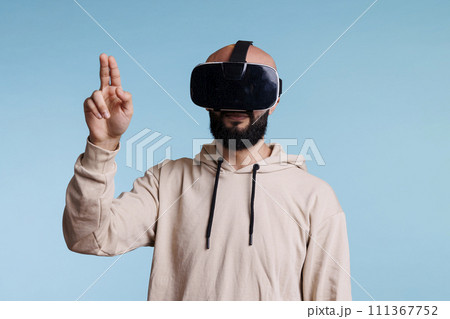 Young man in vr helmet making tap gesture with two fingers while exploring metaverse. Person in headset enjoying simulation activity and playing virtual reality games in cyberspace Young man in vr helmet making tap gesture with two fingers while exploring metaverse. Person in headset enjoying simulation activity and playing virtual reality games in cyberspace 111367752
