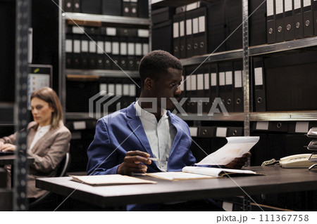 African american bookkeeper analyzing management files, searching for administrative report in corporate repository. Businessman working late at night at bookkeeping record in storage room African american bookkeeper analyzing management files, searching for administrative report in corporate repository. Businessman working late at night at bookkeeping record in storage room 111367758