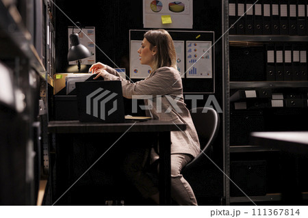 Bookkeeper analyzing bureaucracy record, working overtime at management research in arhive room. Businesswoman in formal suit checking accountancy report, searching for administrative files Bookkeeper analyzing bureaucracy record, working overtime at management research in arhive room. Businesswoman in formal suit checking accountancy report, searching for administrative files 111367814