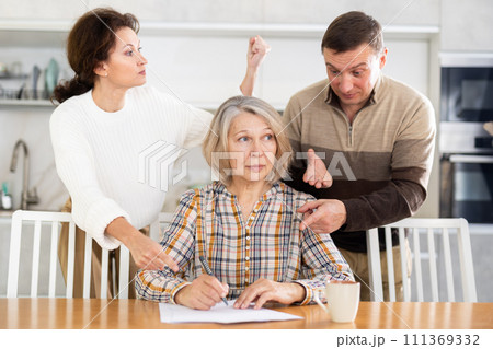 Old woman compiling heritage document while middle-aged man and woman competing in the kitchen 111369332