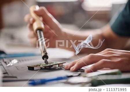 Hands of unrecognizable caucasian electronic repairman soldering mainboard sitting at workshop table 111371995