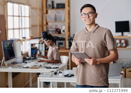 Male workshop owner confidently posing at workplace, black female technician working on background Male workshop owner confidently posing at workplace, black female technician working on background 111371999