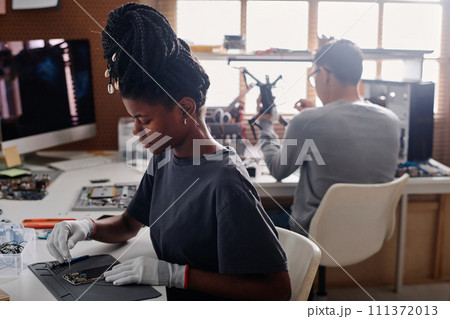 Black woman with braids repairing cellphone at workshop desk, her coworker sitting on background 111372013