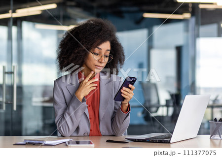 Focused woman with curly hair using smartphone while working in a contemporary office setting, showcasing multitasking and efficiency. Focused woman with curly hair using smartphone while working in a contemporary office setting, showcasing multitasking and efficiency. 111377075
