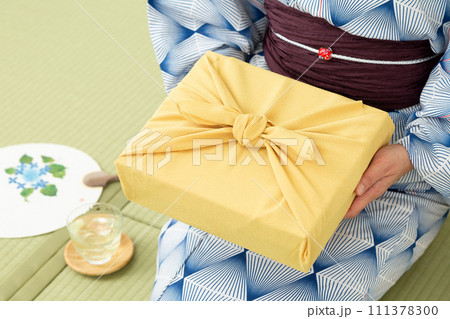 和室の畳に座ってお中元の風呂敷包みを持つ浴衣姿のミドル女性 111378300