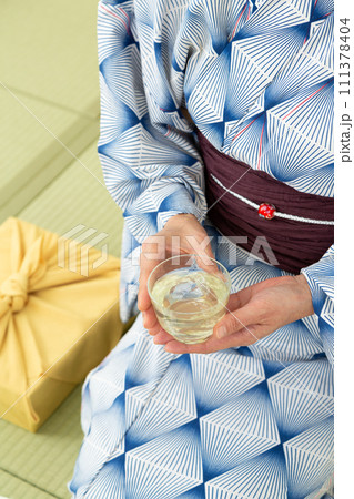 和室の畳に座って冷茶を飲む浴衣姿のミドル女性とお中元の風呂敷包み 111378404