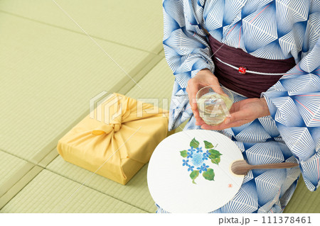 和室の畳に座って冷茶を飲む浴衣姿のミドル女性とお中元の風呂敷包み 111378461