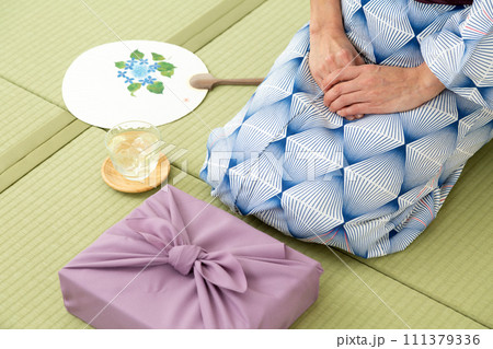 和室の畳に座る浴衣姿のミドル女性とお中元の風呂敷包み 和室の畳に座る浴衣姿のミドル女性とお中元の風呂敷包み 111379336
