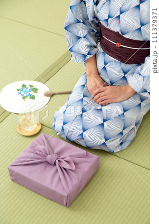 和室の畳に座る浴衣姿のミドル女性とお中元の風呂敷包み 111379371