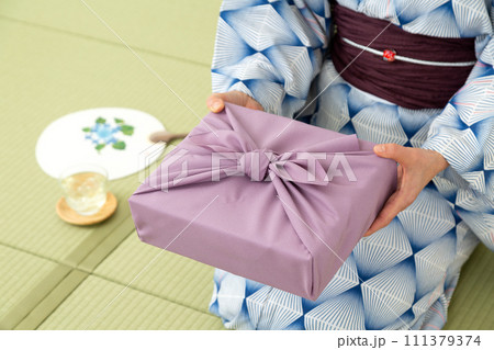 和室の畳に座ってお中元の風呂敷包みを持つ浴衣姿のミドル女性 111379374