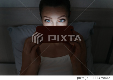 Young woman laying on bed and addictively staring at her smartphone late at night. 111379682