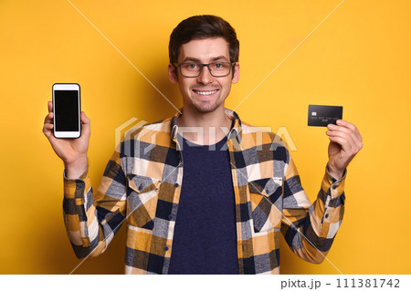Optimistic guy holding smartphone and plastic credit card isolated over yellow background 111381742