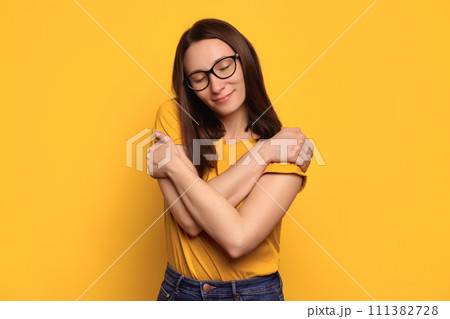 Pretty caucasian woman in eyeglasses hugging herself isolated on yellow studio wall Pretty caucasian woman in eyeglasses hugging herself isolated on yellow studio wall 111382728
