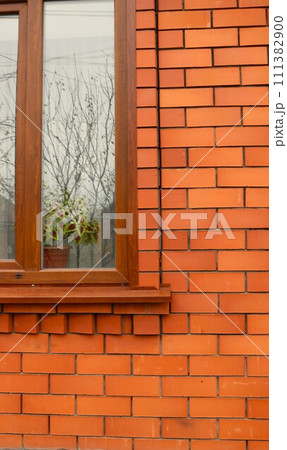 The vertical background of the brick wall of a new residential building with a fragment of a window in a brown plastic frame with a flower on a window and decorating trees on the glass, The vertical background of the brick wall of a new residential building with a fragment of a window in a brown plastic frame with a flower on a window and decorating trees on the glass, 111382900