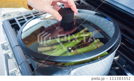 In a modern white kitchen, large organic asparagus is being steamed in a sizable cooking pot. 111383352