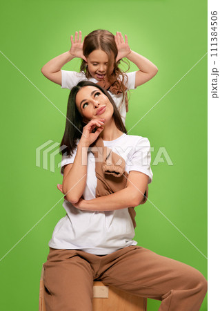 Grimacing and playing. Happy little kid, daughter playing wit her beautiful caring young mother against green studio background Grimacing and playing. Happy little kid, daughter playing wit her beautiful caring young mother against green studio background 111384506
