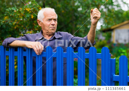 Old elderly farmer stands at fence of his country house and threatens someone with his fist Old elderly farmer stands at fence of his country house and threatens someone with his fist 111385661