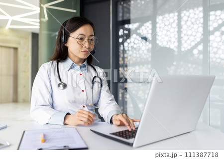 Experienced Asian doctor offers telemedicine services, remotely consulting patients from a clinic office with a headset and laptop. 111387718