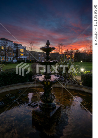 Fountain and pond in a park at sunset with a pink sky. Dusk view in portrait orientation with buildings and trees behind. Fountain and pond in a park at sunset with a pink sky. Dusk view in portrait orientation with buildings and trees behind. 111389500