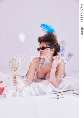 Beautiful young woman with feather head accessories and sunglasses lying on bed in vintage lingerie and looking on antique mirror Beautiful young woman with feather head accessories and sunglasses lying on bed in vintage lingerie and looking on antique mirror 111389754