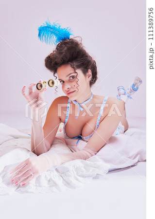 Attractive young woman with opera glasses in vintage lingerie lying on bed against pastel background. Elegant boudoir aesthetics Attractive young woman with opera glasses in vintage lingerie lying on bed against pastel background. Elegant boudoir aesthetics 111389756