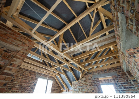 Attic space of a building under construction with wooden beams of a roof structure and brick walls. Real estate development concept. Attic space of a building under construction with wooden beams of a roof structure and brick walls. Real estate development concept. 111390864