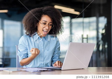 A woman is happily using her laptop computer while sitting at a desk in a room with fun decor and a smile on her face. A woman is happily using her laptop computer while sitting at a desk in a room with fun decor and a smile on her face. 111391208