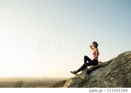 Woman hiker sitting on a steep big rock enjoying warm summer day. Young female climber resting during sports activity in nature. Active recreation in nature concept. Woman hiker sitting on a steep big rock enjoying warm summer day. Young female climber resting during sports activity in nature. Active recreation in nature concept. 111391704