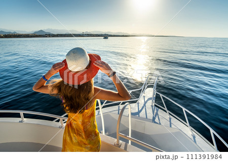 Young woman with long hair wearing yellow dress and straw hat standing on white yacht deck enjoying view of blue sea water. 111391984