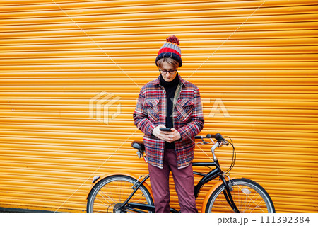Young hipster man using mobile phone while sitting on cruiser bicycle near orange wall. Student using 5g network,rests after riding. Eco friendly transport for carbon footprint. Sustainable lifestyle 111392384