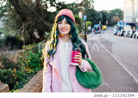 Stylish young smiling hipster woman with color hair walking on street in pink outfit with reusable coffee cup wearing coat, knitted hat, fur bag, happy mood, seasonal fashion, Barbiecore style trend Stylish young smiling hipster woman with color hair walking on street in pink outfit with reusable coffee cup wearing coat, knitted hat, fur bag, happy mood, seasonal fashion, Barbiecore style trend 111392394