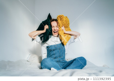 frustrated woman covers ears with pillows, screaming feels furious exhausted exasperated sitting on bed. mad irritated millennial suffers from head ache lack of self-control. Noise and stress concept. 111392395